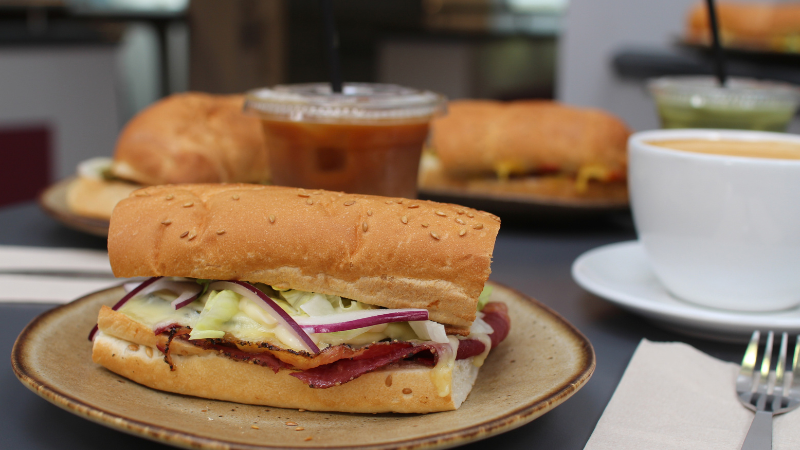 A sub sandwich filled with meat, cheese, lettuce, and red onion on a plate, surrounded by other subs, iced drinks, and a cup of coffee.
